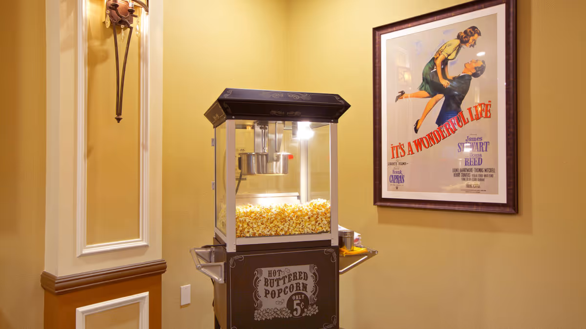 A vintage popcorn machine filled with popcorn sits against a yellow wall beside a framed "It's a Wonderful Life" movie poster.