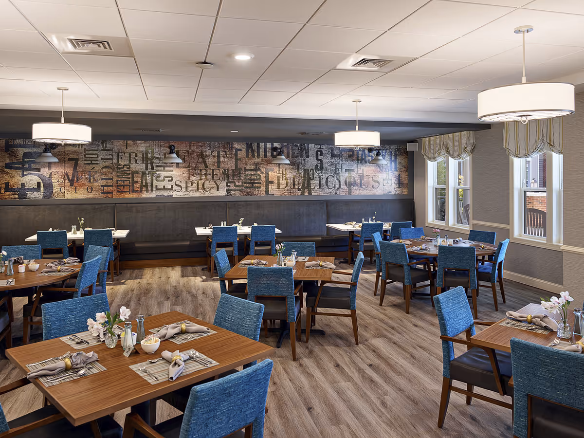 A well-lit dining room with multiple wooden tables set with placemats, napkins, and small flower arrangements. Blue upholstered chairs surround the tables. The room has large windows with striped valances, modern pendant lights hanging from the ceiling, and a decorative wall with various words related to food and dining.