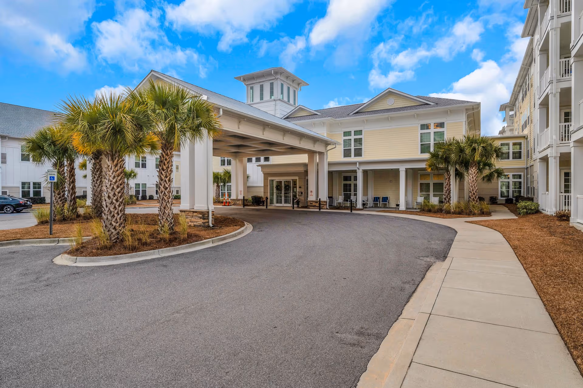 Exterior view of The Barclay at Little River senior living facility showing the main entrance with a covered drop-off area, palm trees, a curved driveway, and a clear blue sky with some clouds.