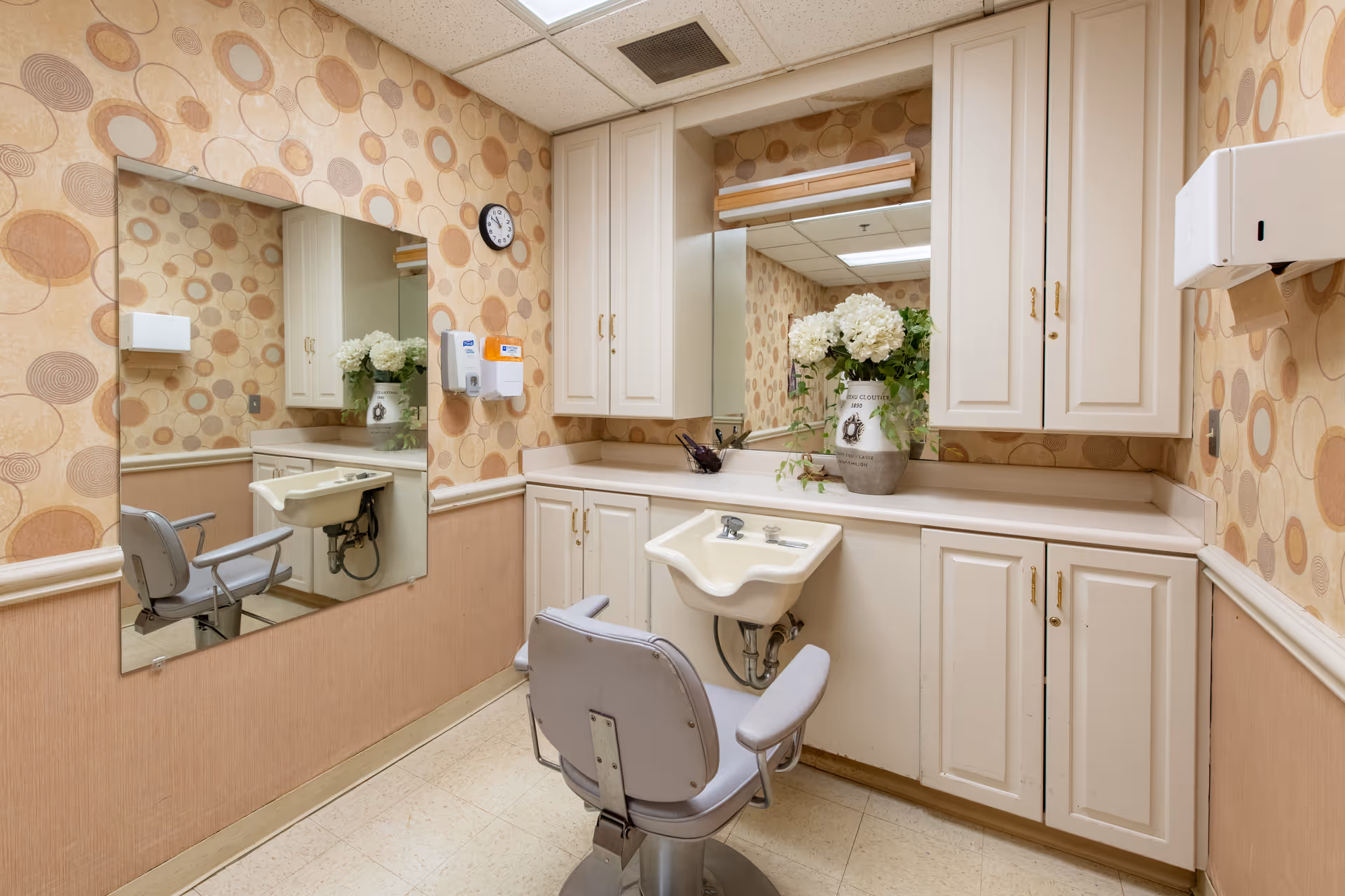 Interior view of a small salon or grooming area in a senior living facility. The room features a single salon chair in front of a small sink with cabinets above and below the countertop. The walls have a patterned wallpaper with circular designs in shades of beige and brown. A large mirror is mounted on one wall, reflecting the room. A clock, a soap dispenser, and a paper towel dispenser are also visible on the walls. A vase with white flowers and green leaves sits on the countertop beneath another mirror.