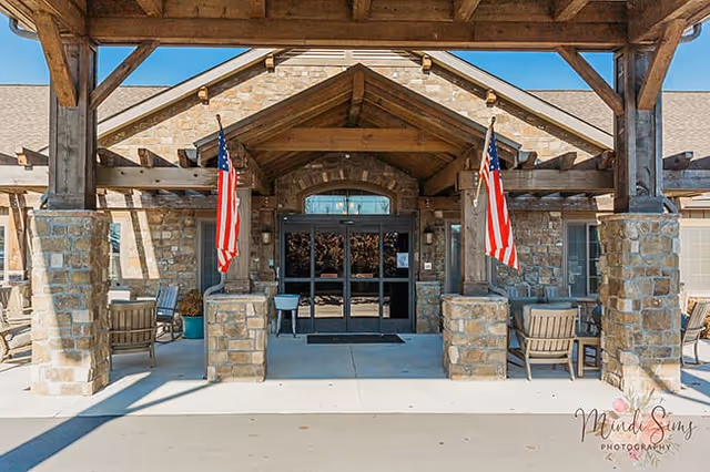 Entrance of a senior living facility with a stone facade and wooden beams supporting a covered porch. Two American flags hang on either side of the entrance, and there are outdoor chairs and tables arranged on both sides under the porch.