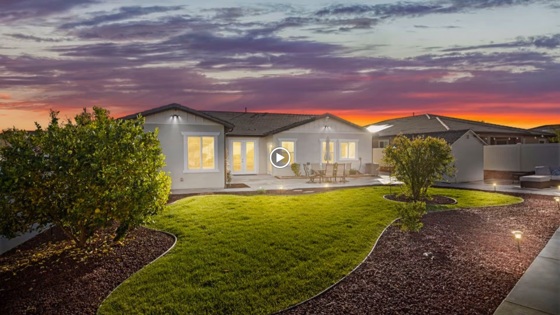 Exterior view of a single-story assisted living facility with a landscaped yard, patio seating, and a sunset sky.