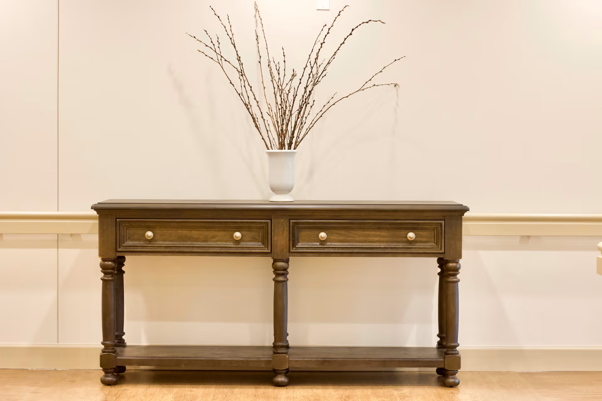A wooden console table with two drawers and turned legs against a beige wall. On top of the table is a white vase holding tall, thin branches. The floor is light-colored wood.