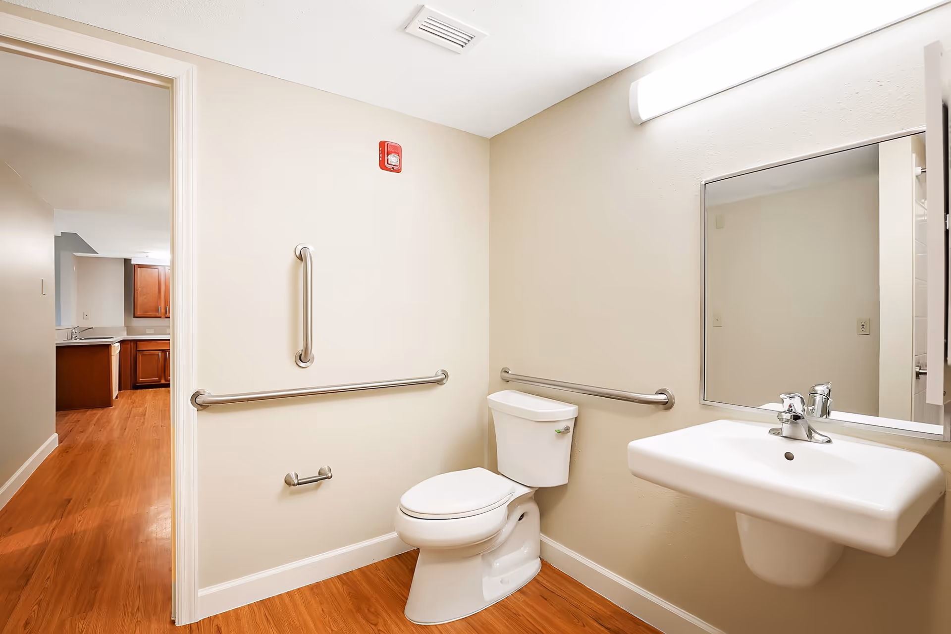A clean, accessible bathroom with a white toilet, a wall-mounted sink, a large mirror above the sink, and stainless steel grab bars on the walls. The floor has wood-like flooring, and the bathroom opens into a hallway with a view of a kitchen area with wooden cabinets.