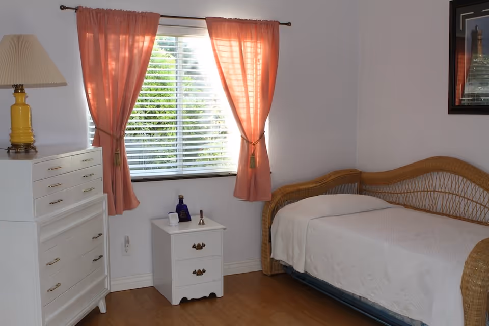 A small bedroom with a wicker daybed covered with a white blanket, a white chest of drawers with a yellow lamp on top, a white nightstand with decorative items, and a window with pink curtains tied back, letting in natural light.