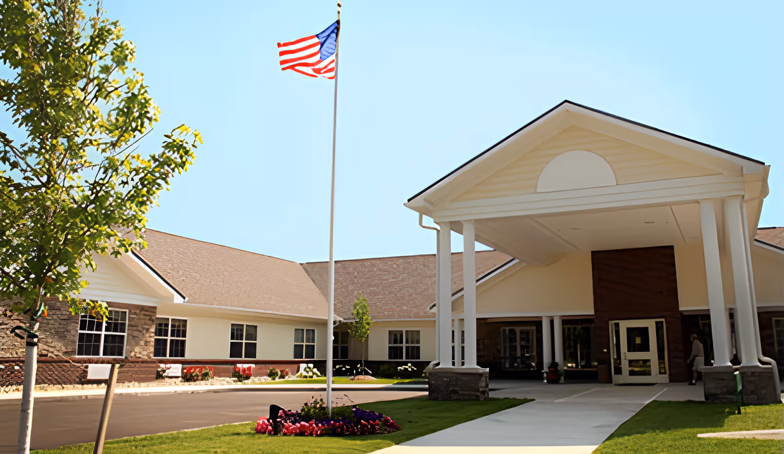 Exterior view of The Willows at Okemos facility showing the entrance with a covered driveway supported by white columns, an American flag on a flagpole, a tree, and a flower bed in front of the building under a clear blue sky.