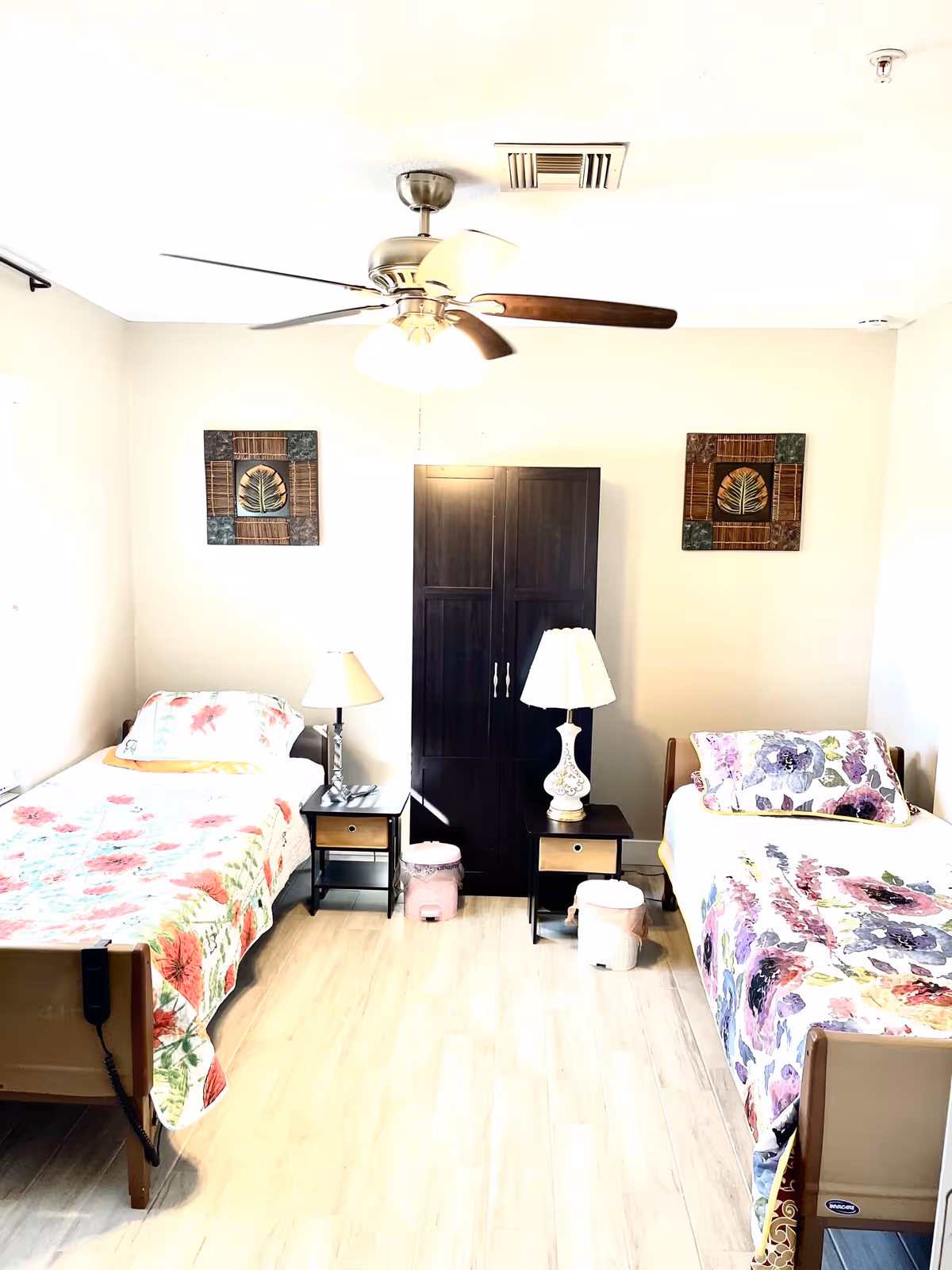 A bright room with two single beds, each with floral bedspreads. Between the beds are two small nightstands with lamps and a dark wooden wardrobe in the center. The room has light-colored wooden flooring, a ceiling fan with lights, and two framed leaf-themed artworks on the wall.