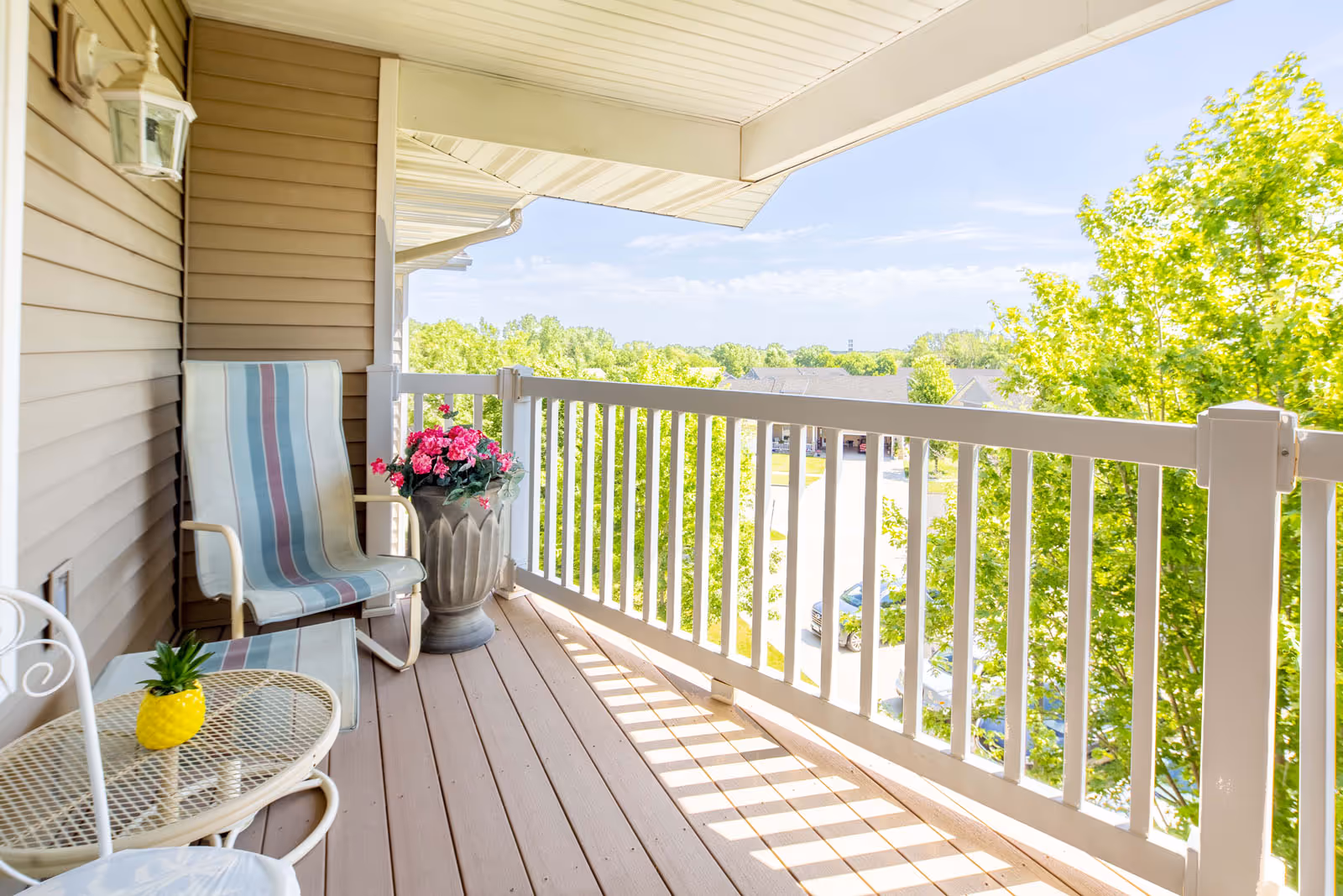 Sunny balcony with a striped chair, small table, and potted flowers overlooking trees and a parking area.