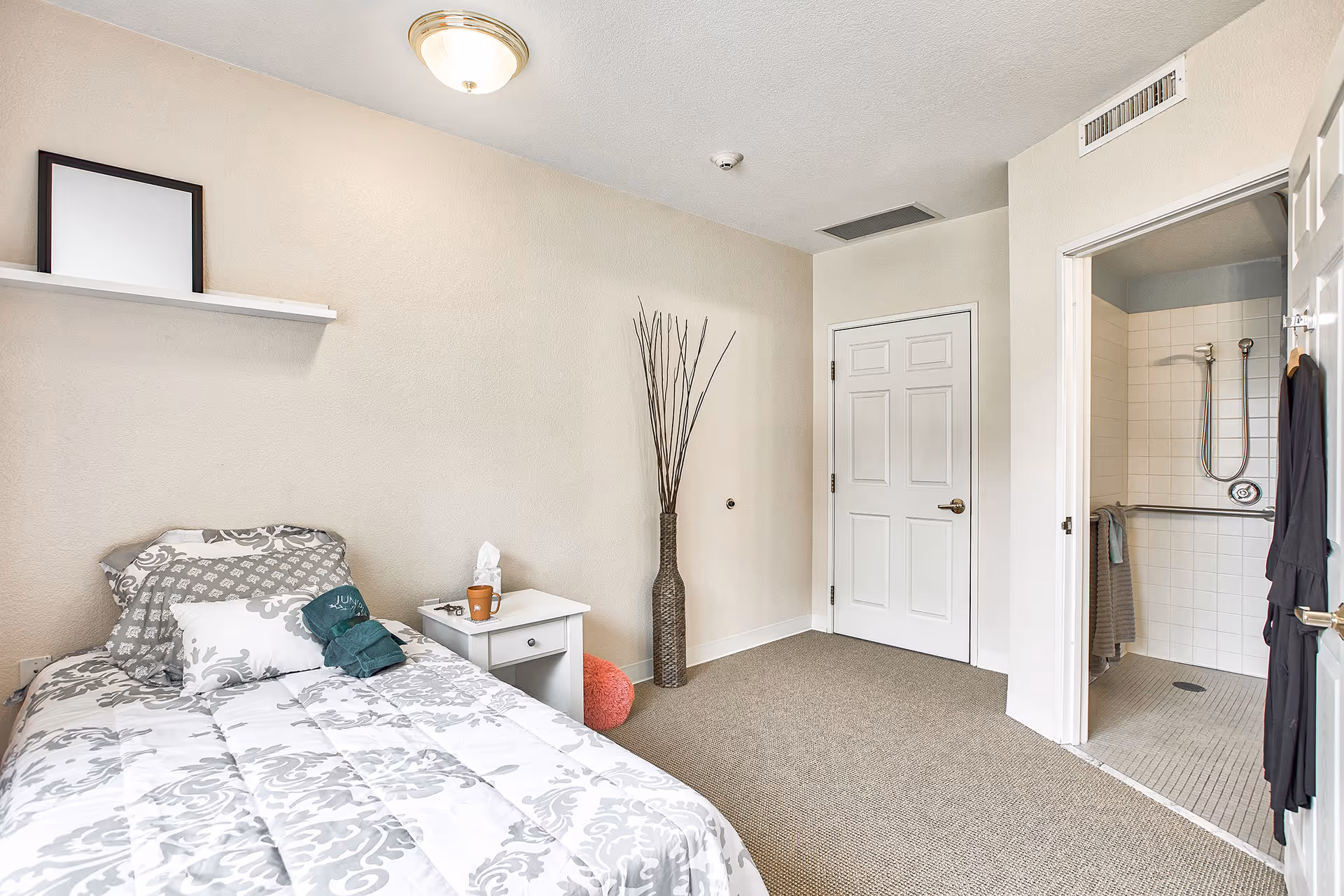 A small bedroom with a single bed covered in a white and gray patterned comforter and pillows. Next to the bed is a white nightstand with a tissue box, a cup, and some small items. A tall decorative vase with long sticks is placed against the wall. There is a door leading to a bathroom with a walk-in shower that has a grab bar and a handheld showerhead. The room has beige walls and carpeted floor.
