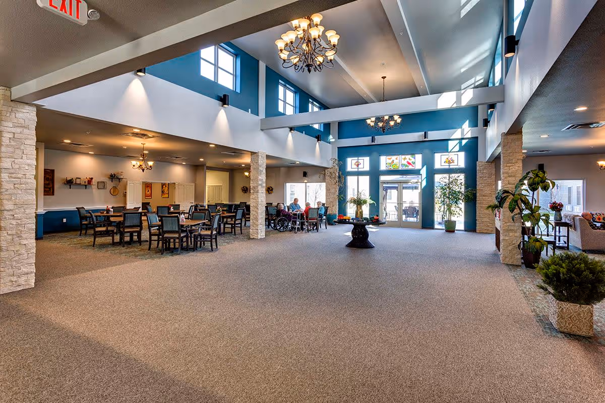 Spacious and well-lit common area in an assisted living facility with high ceilings, large windows, and chandeliers. The room features a dining area with tables and chairs, a seating area with sofas, and several plants. A group of elderly people are seated and conversing near the dining area. The walls are painted blue and white, and there is an exit sign visible on the ceiling.