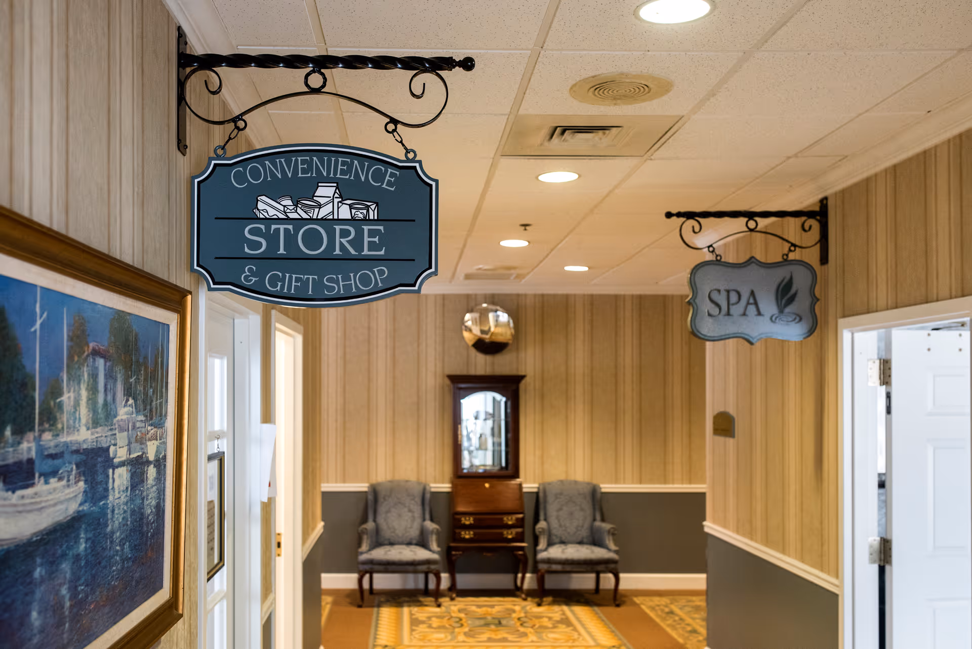 Interior hallway of a senior living facility with signs hanging from the ceiling indicating directions to a convenience store & gift shop and a spa. The hallway has beige and gray walls, a patterned carpet, two upholstered chairs, a wooden cabinet with a mirror, and a framed painting on the left wall.
