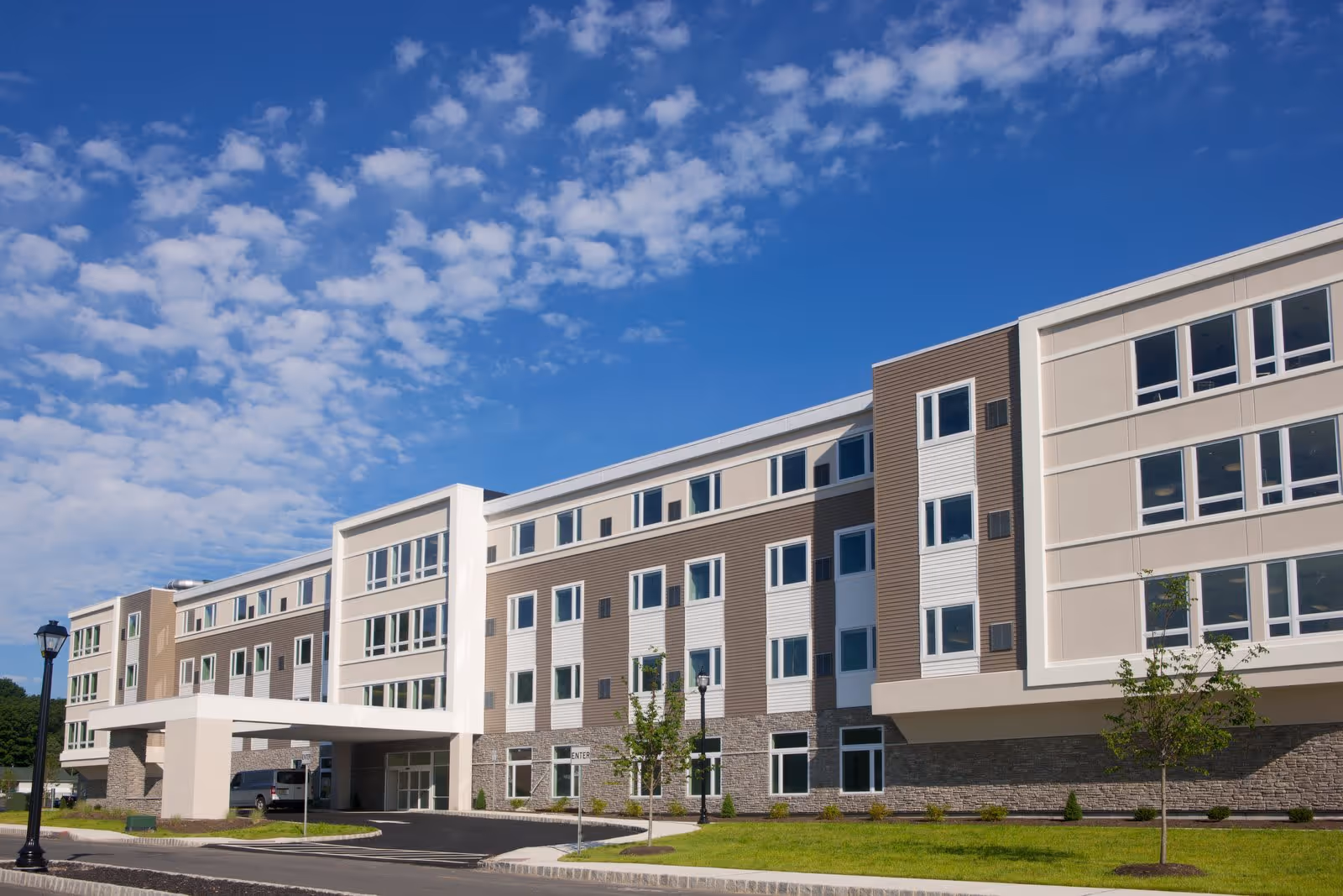 Exterior view of a modern multi-story senior living facility building under a blue sky with scattered clouds. The building features large windows, a covered entrance, and landscaped grounds with small trees and grass.
