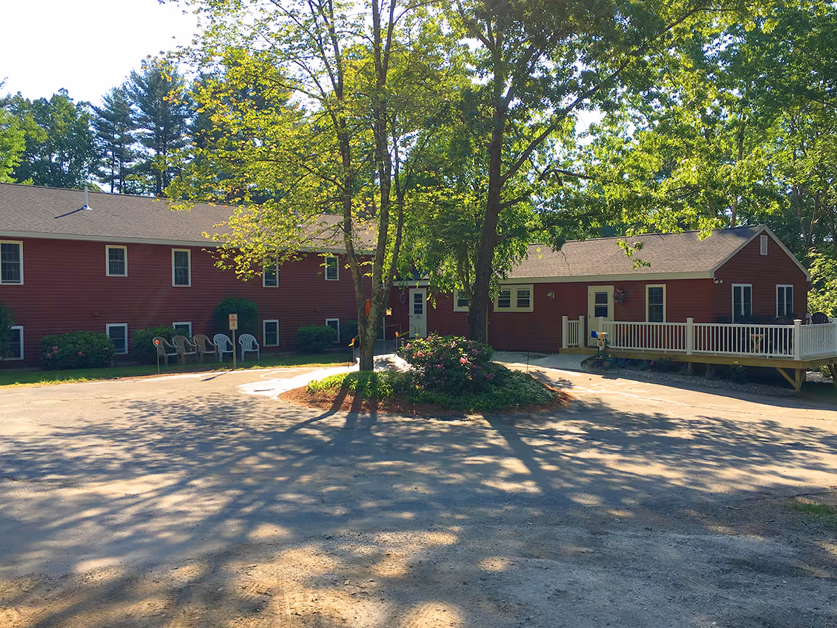 Exterior view of a red building with white trim surrounded by trees and greenery. There is a circular driveway with a landscaped island in the center featuring bushes and flowers. Several chairs are placed near the building, and a wooden deck with a railing is visible on the right side.