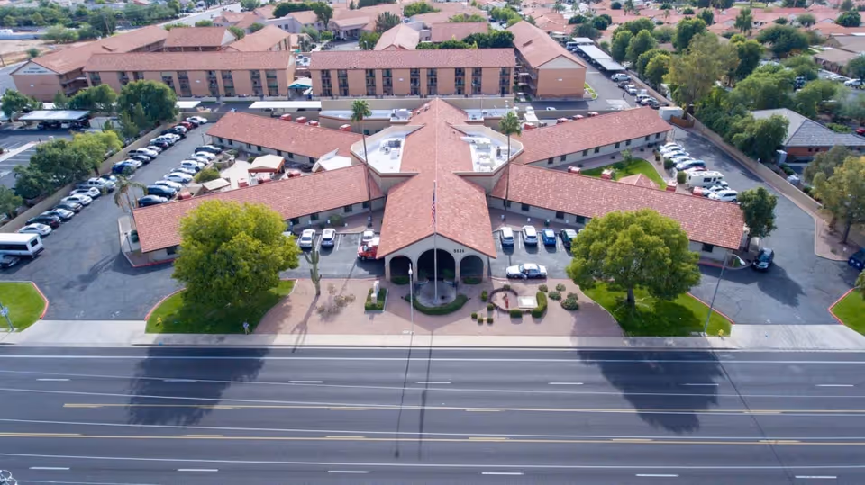 Aerial view of a large facility with a star-shaped layout and red-tiled roofs, surrounded by parking lots and greenery. The building is located next to a multi-lane road, with residential houses visible in the background.