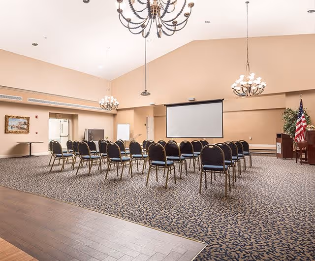 A large meeting or conference room with rows of chairs facing a blank projector screen. The room features beige walls, patterned carpet, three chandeliers hanging from a high ceiling, an American flag near a podium, a television, and a framed picture on the wall.