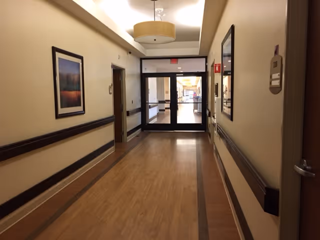 A well-lit hallway in a senior living facility with wooden flooring, beige walls, handrails on both sides, framed artwork on the walls, and double glass doors at the end.