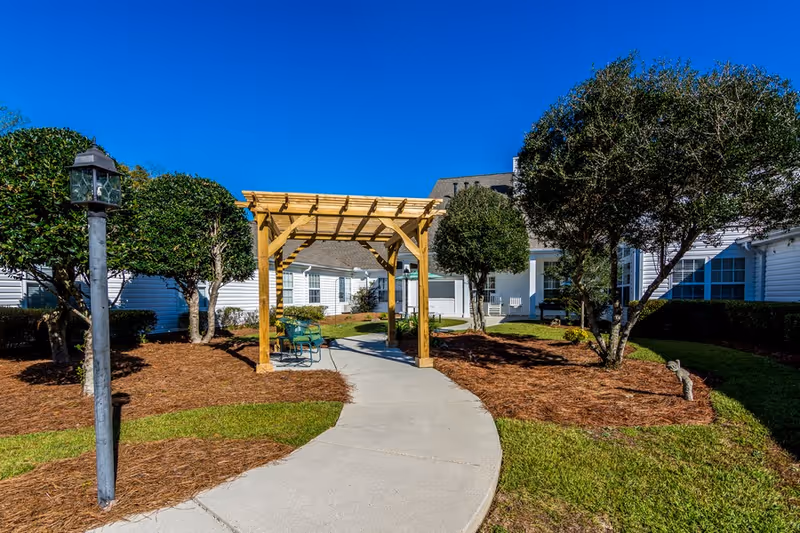 Outdoor garden area at The Pines at Florence Assisted Living & Memory Care featuring a curved concrete walkway, a wooden pergola with benches underneath, neatly trimmed trees and bushes, and a clear blue sky.