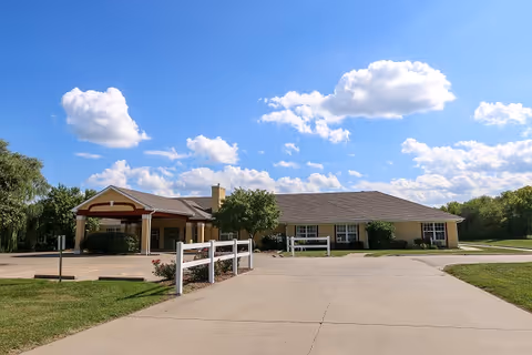 Single-story building with a covered entrance, surrounded by green grass and trees under a blue sky with scattered clouds. The building appears to be a senior living facility with a paved driveway and white fencing near the entrance.