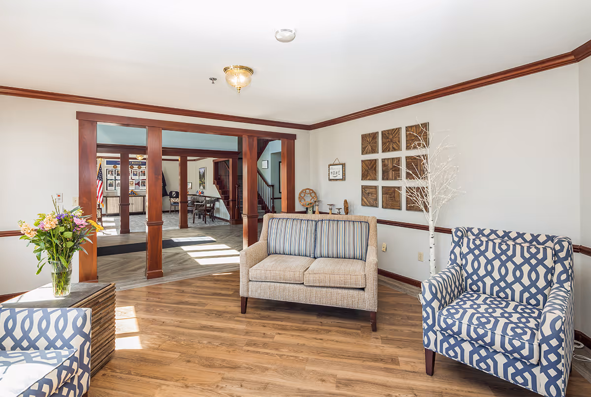 A bright and inviting living room area with wooden flooring and white walls accented by dark wood trim. The room features a beige loveseat with striped cushions, a blue and white patterned armchair, a small side table with a vase of colorful flowers, and decorative wall art. In the background, there is an open doorway with wooden columns leading to another room with more seating and a staircase.