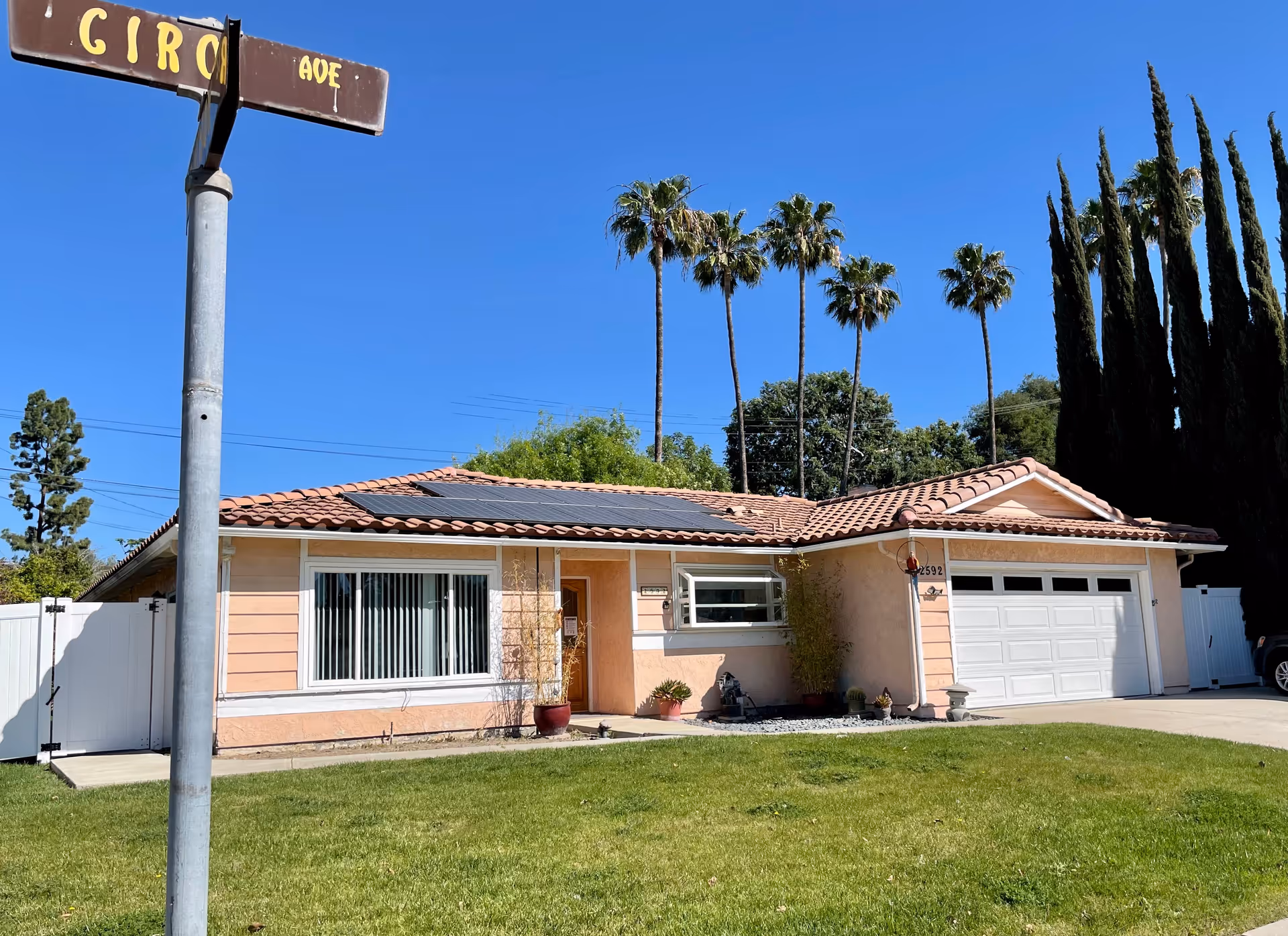 Single-story peach-colored house with a tiled roof and solar panels, a white garage door, and a green lawn in front. There are tall palm trees and other trees in the background under a clear blue sky. A street sign is visible on the left side of the image.