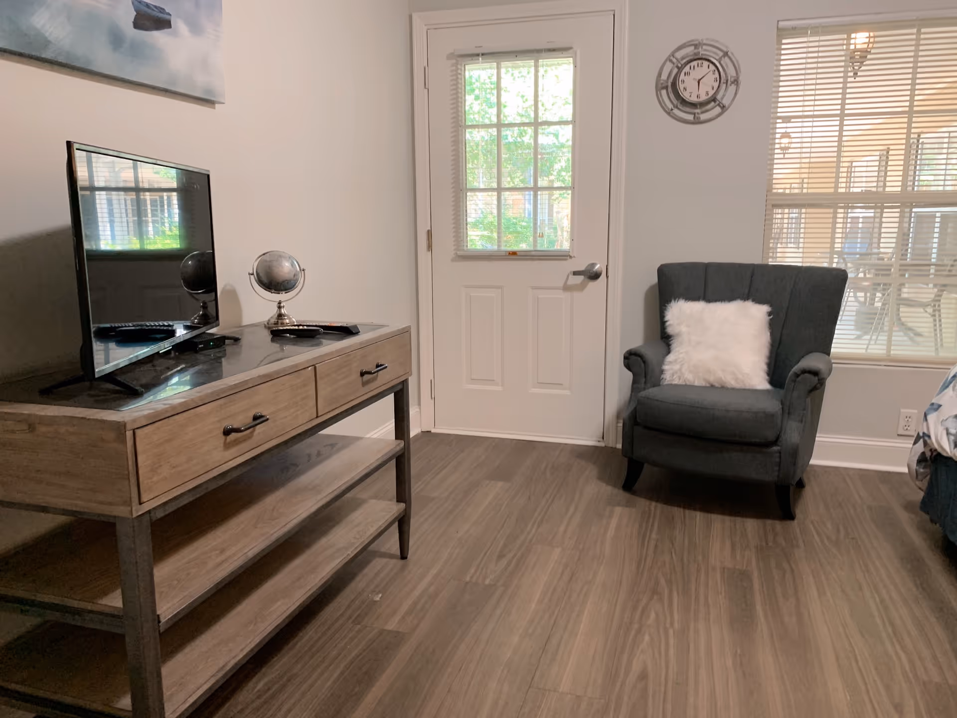 Small furnished room with a TV on a wooden console, a gray armchair with a white pillow, and a door and window.