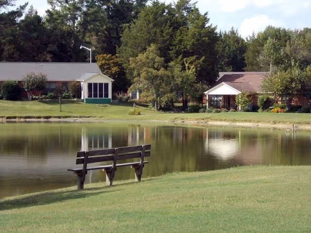 A wooden bench beside a calm pond with grassy shore and small houses and trees reflected in the water.