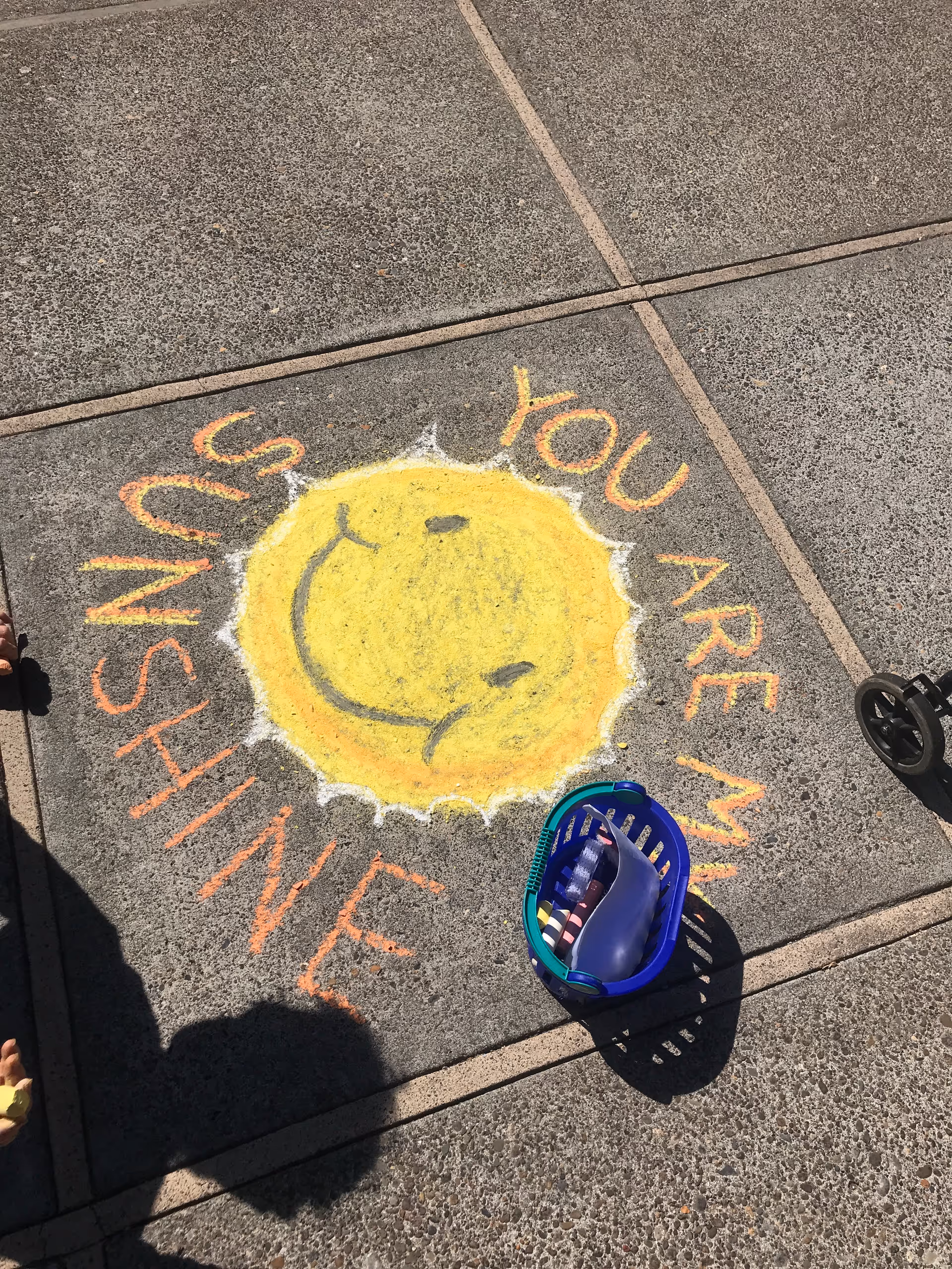Sidewalk chalk drawing of a smiling yellow sun with the words 'YOU ARE MY SUNSHINE' written around it on a concrete pavement. A blue basket with chalk pieces is placed nearby.
