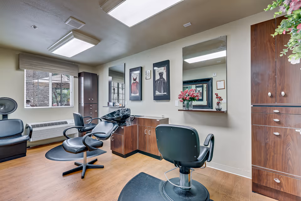 Interior view of a salon area in a senior living facility with salon chairs, a hair washing station, mirrors on the wall, wooden cabinets, and a window with a view outside. There are framed pictures and flower vases on the counter.