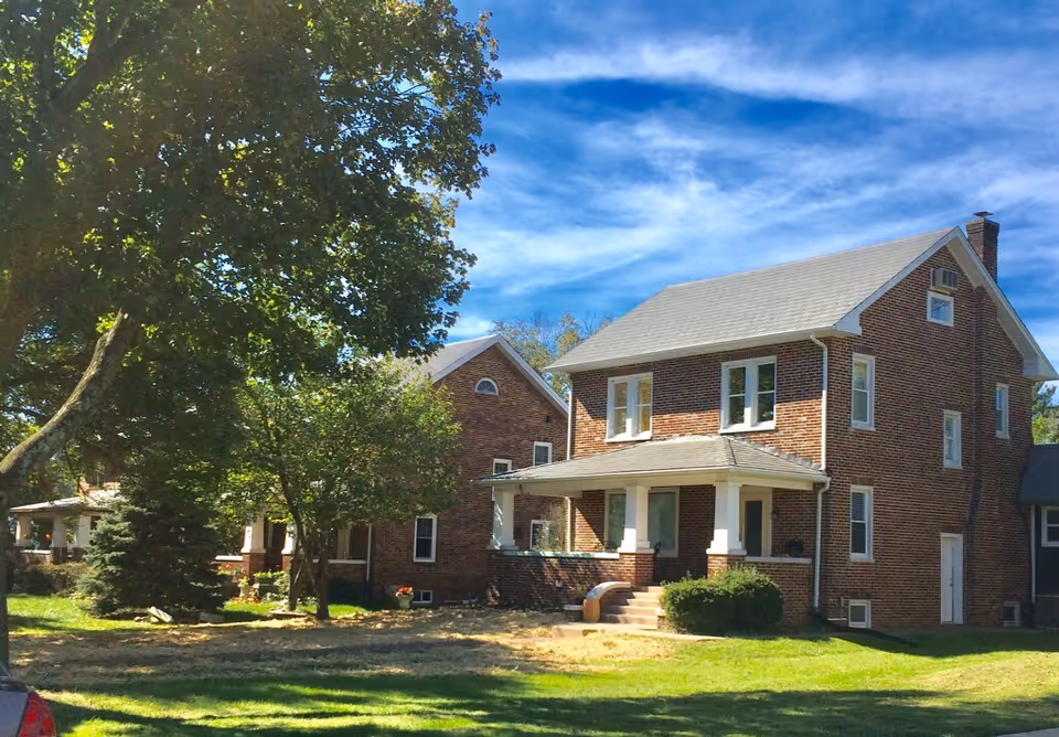 A two-story brick house with a covered front porch, surrounded by green grass and trees under a blue sky with some clouds.