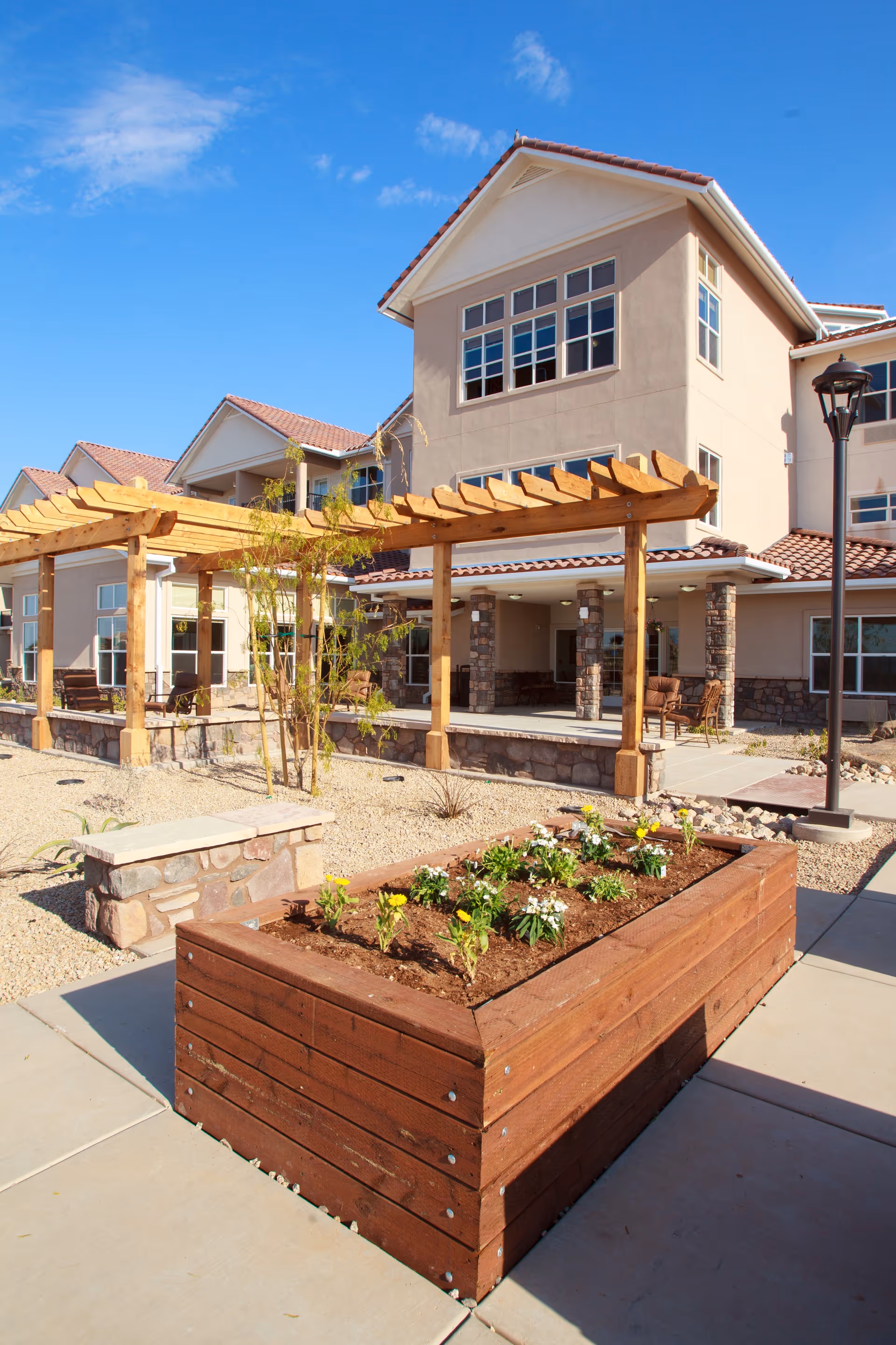 Outdoor area of a senior living facility featuring a raised wooden garden bed with small flowering plants, a wooden pergola, stone benches, and a multi-story building with large windows and a tiled roof under a clear blue sky.