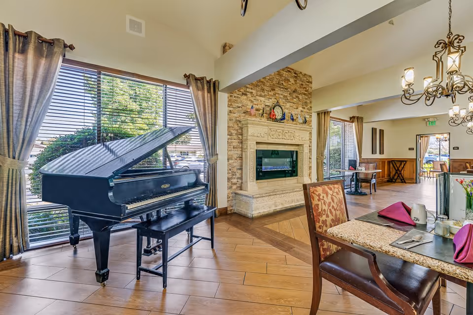 Interior view of a senior living facility dining area featuring a black grand piano near large windows with curtains, a stone fireplace with decorative items on the mantel, and a dining table set with purple napkins, silverware, and cups. The room has wooden flooring and elegant chandeliers hanging from the ceiling.
