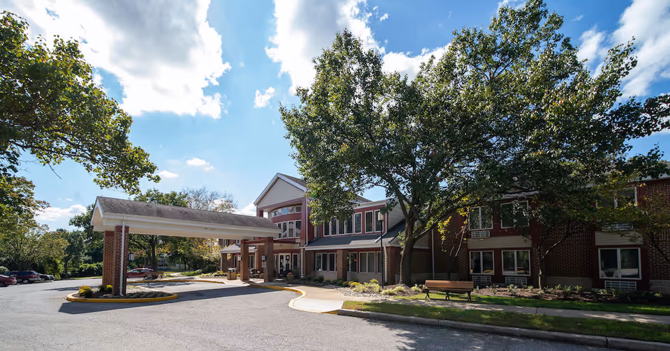 Exterior view of Independence Court of Hyattsville, a two-story brick building with large windows. The entrance features a covered drop-off area supported by brick columns. Trees and landscaping surround the building, and a bench is visible near the sidewalk. The sky is partly cloudy with sunlight illuminating the scene.
