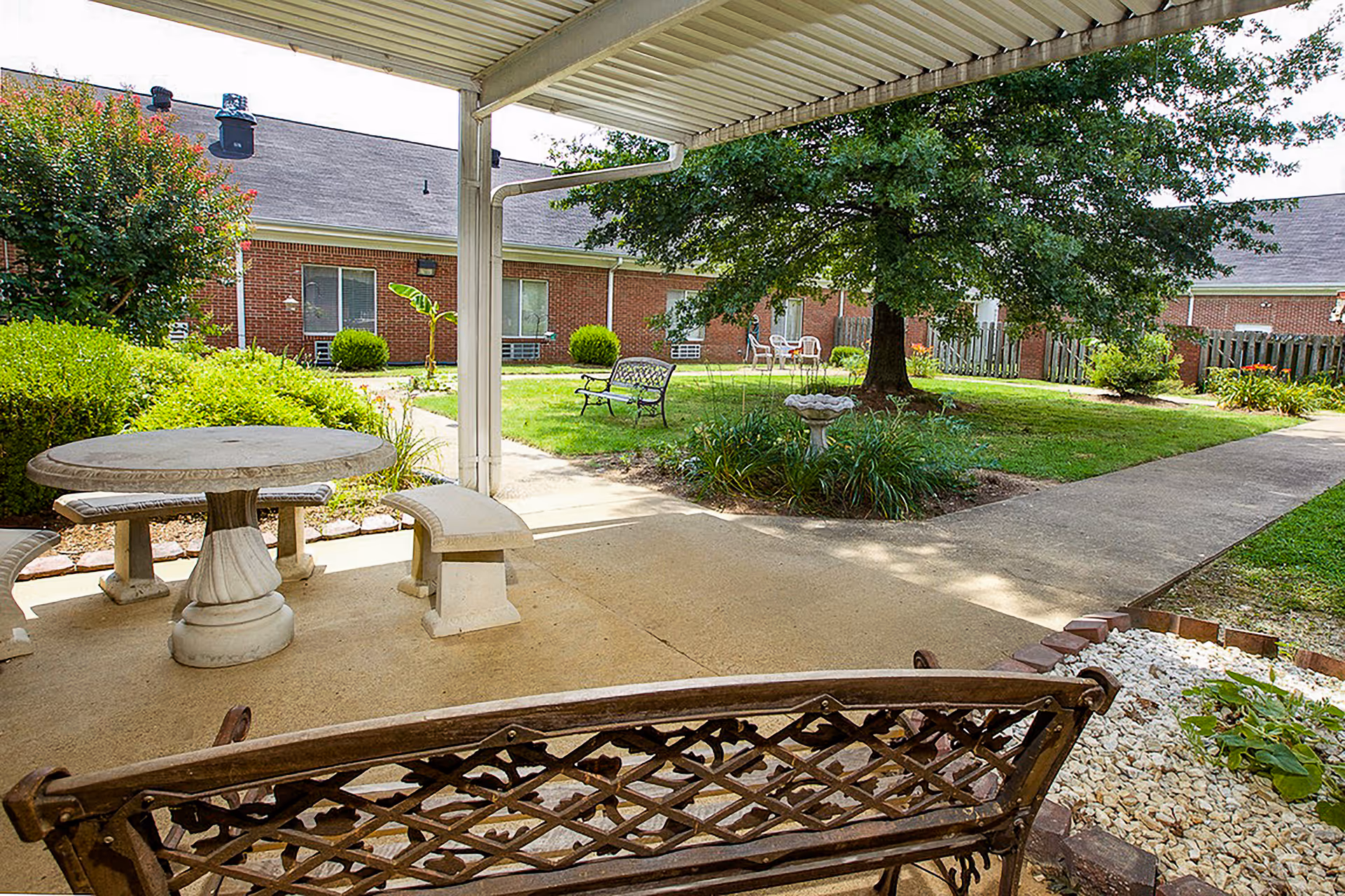 Covered patio area with stone table and benches, overlooking a green courtyard with a large tree, benches, a birdbath, and brick buildings in the background.