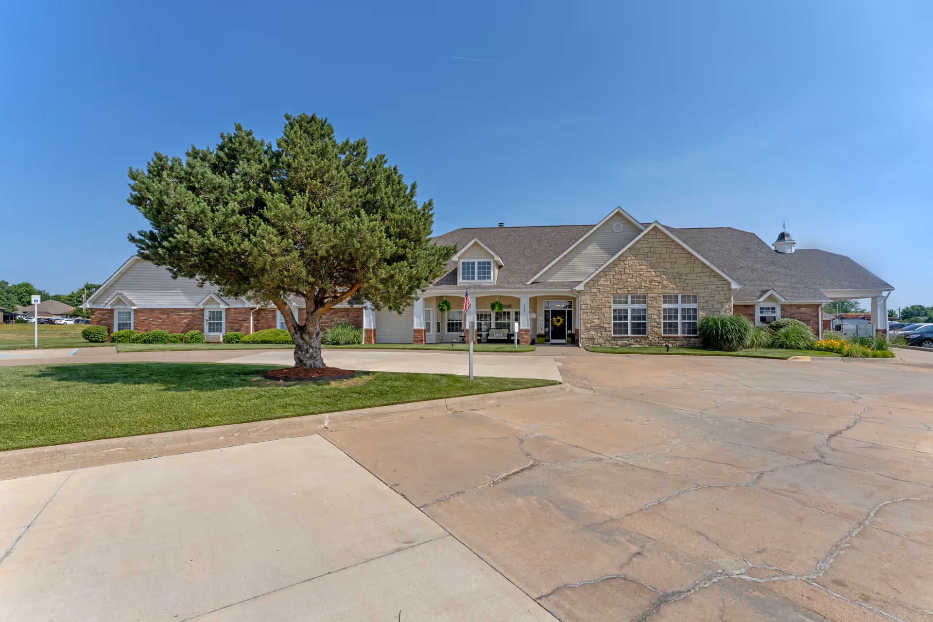 Exterior view of a single-story senior living facility building with a combination of brick and stone facade, a large tree on a grassy area in front, and a clear blue sky overhead.