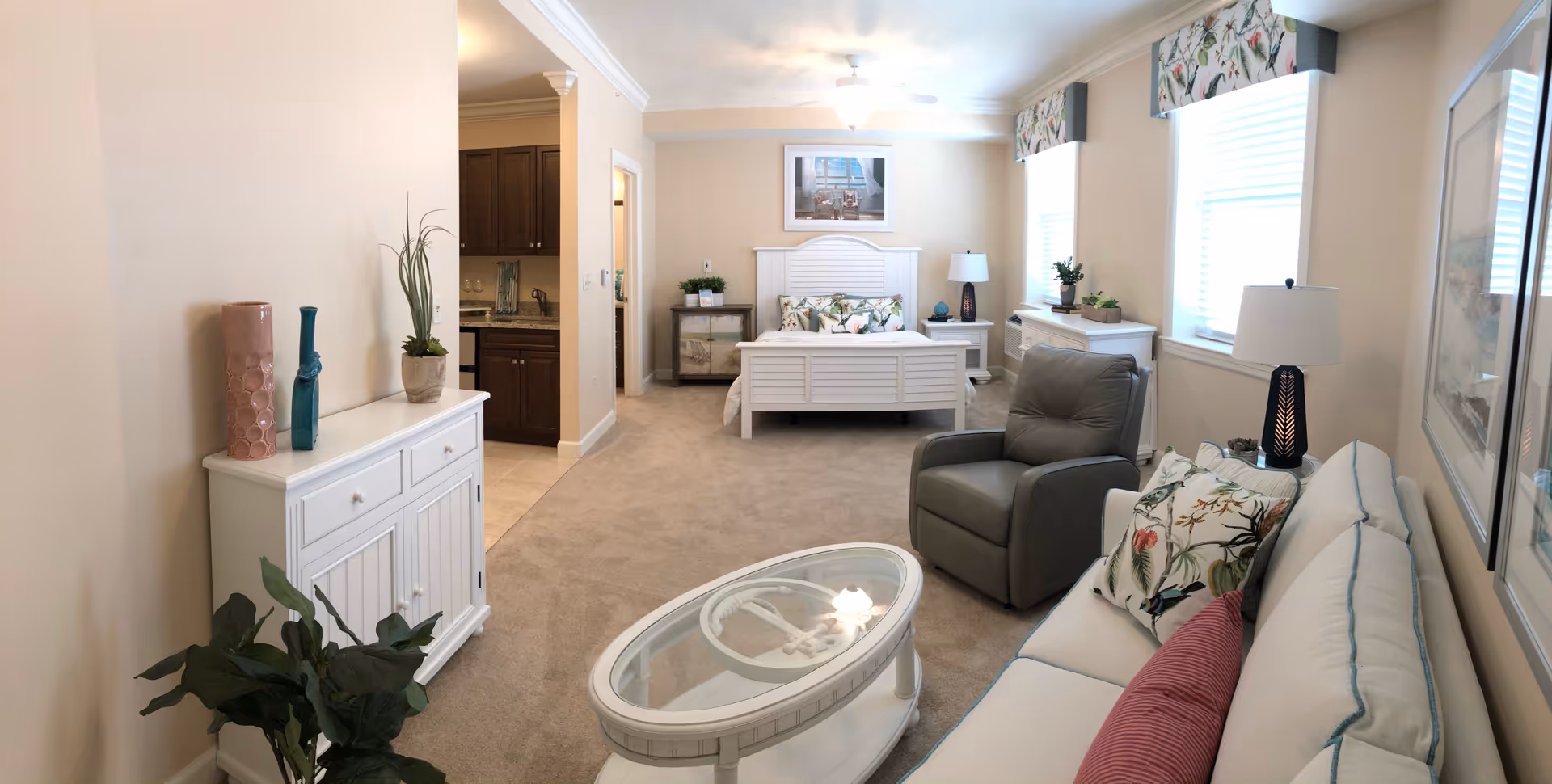 A bright and cozy senior living apartment featuring a white bed with floral pillows, a gray armchair, a white sofa with colorful cushions, an oval glass coffee table, and a white cabinet with decorative vases. The room has beige walls, carpeted floor, two large windows with floral valances, and a ceiling fan with light. A small kitchenette with wooden cabinets is visible in the background.