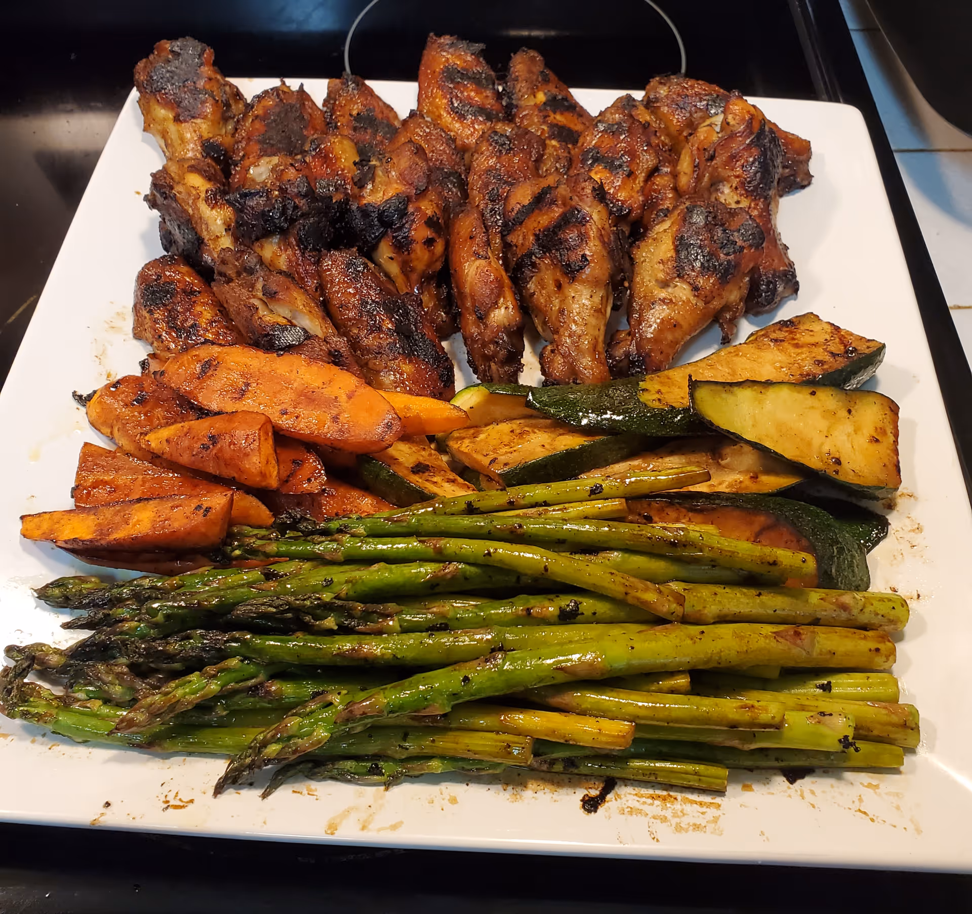 White square plate with grilled chicken drumsticks, roasted asparagus, zucchini, and sweet potato wedges.