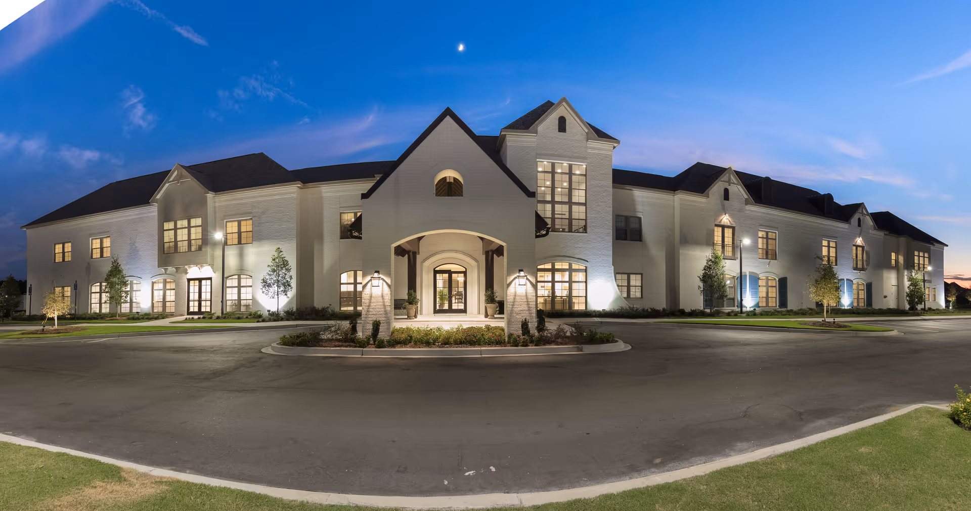 Exterior view of a large, two-story senior living facility building named The Blake at Baxter Village during twilight, with lights illuminating the white brick facade and a clear sky with a crescent moon above.