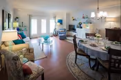 A bright and spacious living and dining area in a retirement community. The living room features a beige sofa, patterned armchair, glass coffee table, and a TV on a stand. The dining area has a round table covered with a gray tablecloth, set with plates and glasses, surrounded by dark wooden chairs. Large windows with white curtains allow natural light to fill the room. The walls are decorated with shelves holding various decorative items and a chandelier hangs above the dining table.