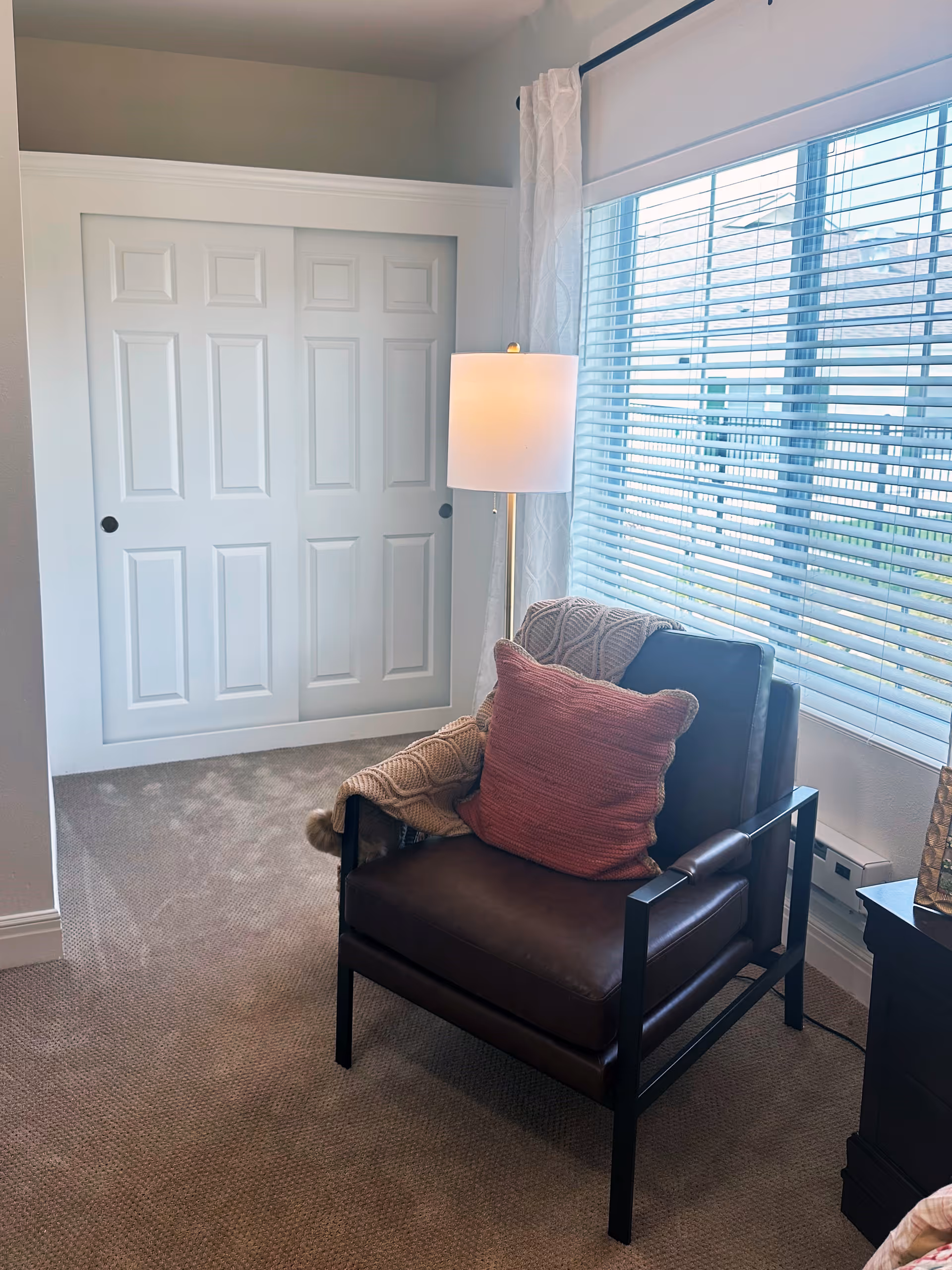 A cozy corner of a room featuring a dark brown armchair with a red cushion and a beige knitted throw draped over the back. Next to the chair is a tall floor lamp with a white lampshade. Behind the chair is a large window with white blinds and sheer white curtains. To the left, there is a white sliding closet door. The floor is carpeted in a neutral tone.