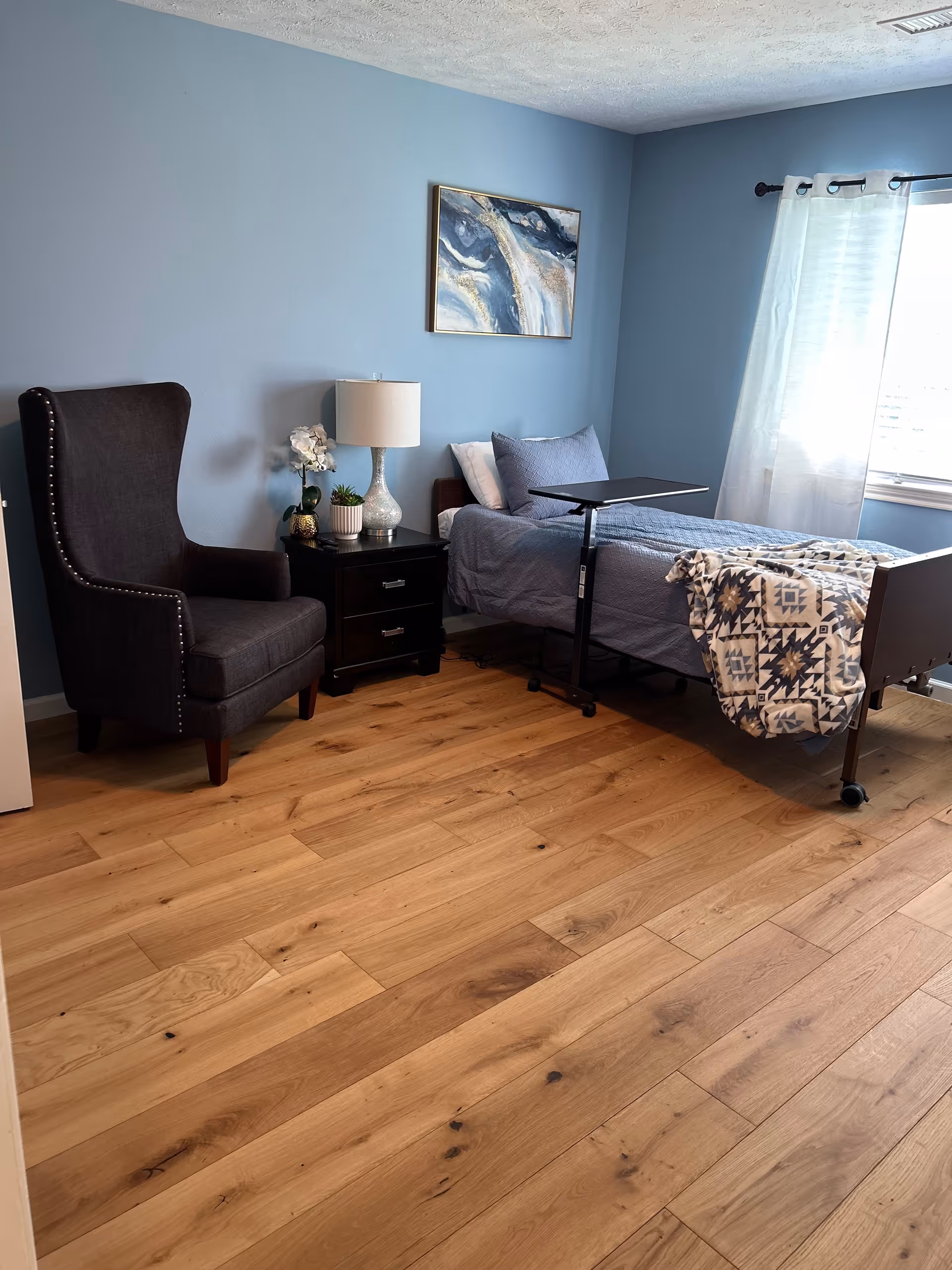A cozy bedroom with light blue walls and wooden flooring. The room features a dark gray upholstered armchair with nailhead trim, a black nightstand with two drawers, a white table lamp, and two small potted plants. There is a single bed with gray bedding, a patterned blanket draped over the foot of the bed, and an adjustable overbed table. A window with white curtains allows natural light to brighten the room. A framed abstract painting hangs on the wall above the nightstand.