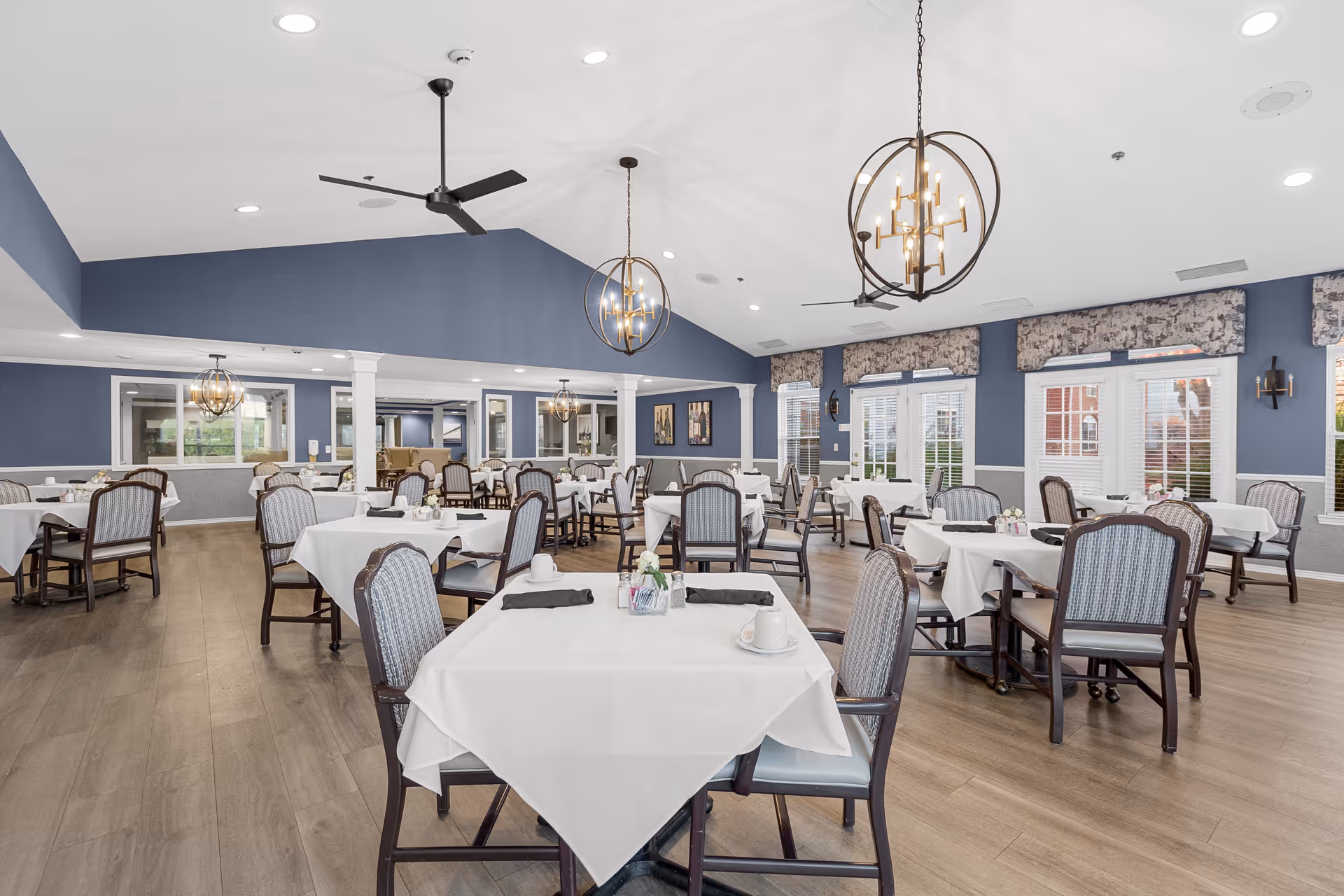 A spacious dining room with multiple tables covered in white tablecloths, each set with cups, napkins, and small flower arrangements. The room features blue walls, large windows with patterned valances, modern chandeliers, ceiling fans, and wood flooring.