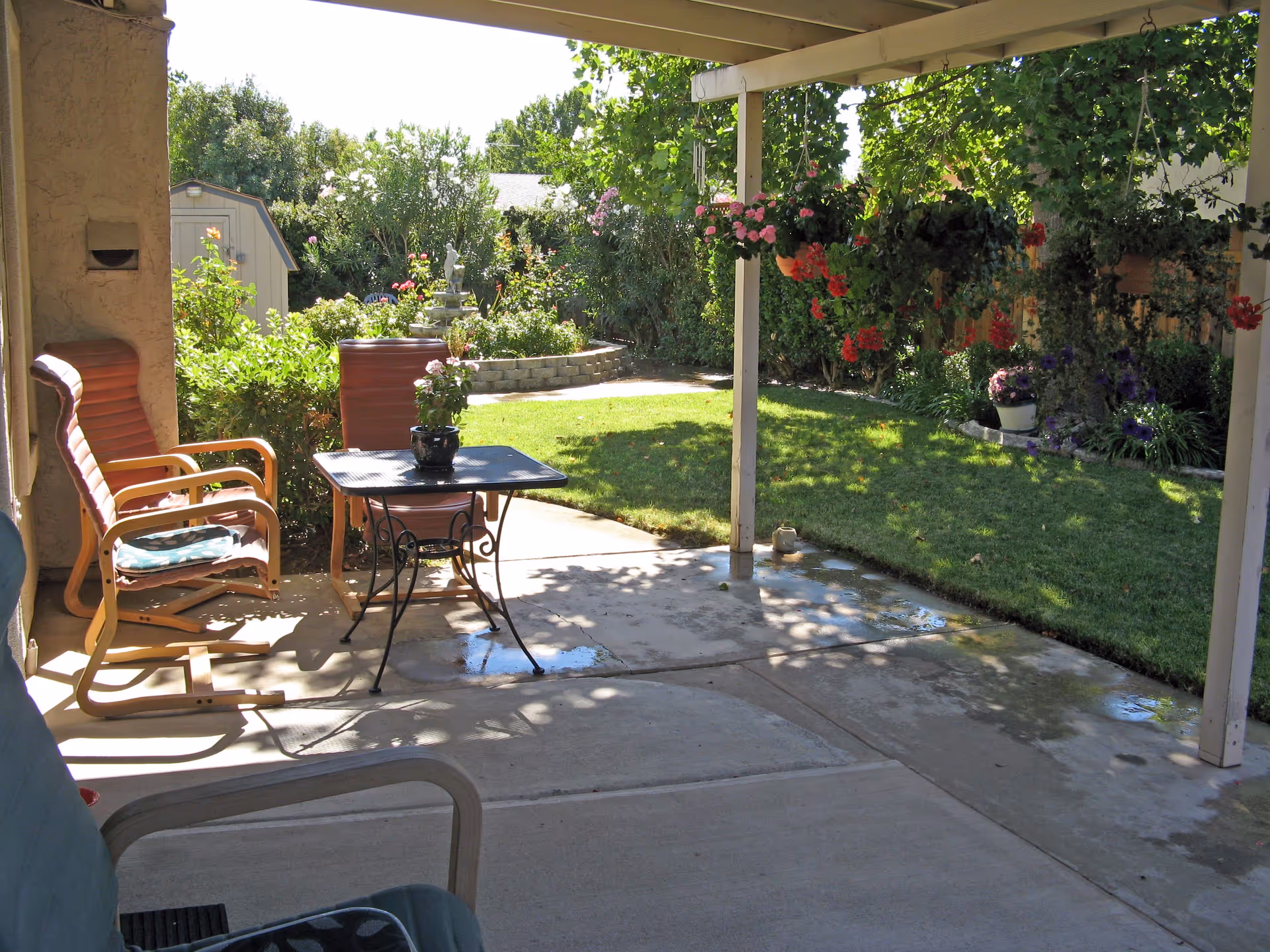 Covered patio with chairs and a small table overlooking a sunlit backyard lawn, garden beds, and hanging flower baskets.
