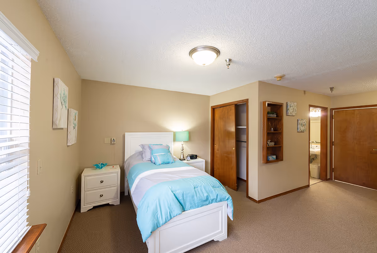 A bright and clean bedroom with a single white bed dressed in light blue and white bedding. There is a white nightstand with a green lamp and a small decorative item. The room has beige walls, a window with white blinds, two framed artworks on the wall, an open closet with wooden sliding doors, and a small built-in wooden shelf with decorative items. A bathroom entrance and a closed wooden door are visible in the background.