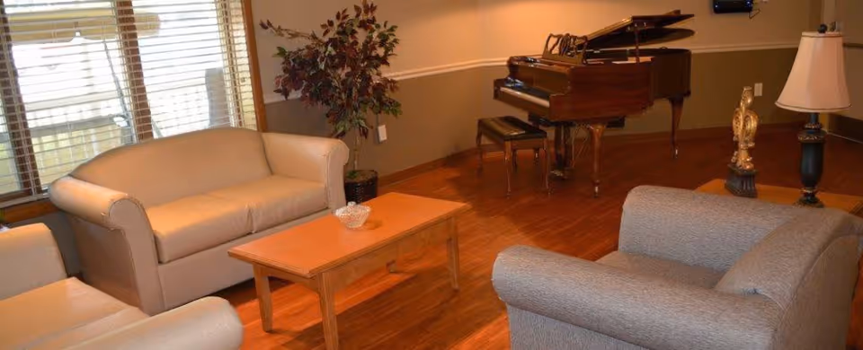 A cozy living room with wooden flooring featuring a beige loveseat, a gray armchair, a wooden coffee table with a decorative bowl, a potted plant near the window with blinds, a grand piano with a bench, and a side table with a lamp and decorative items.
