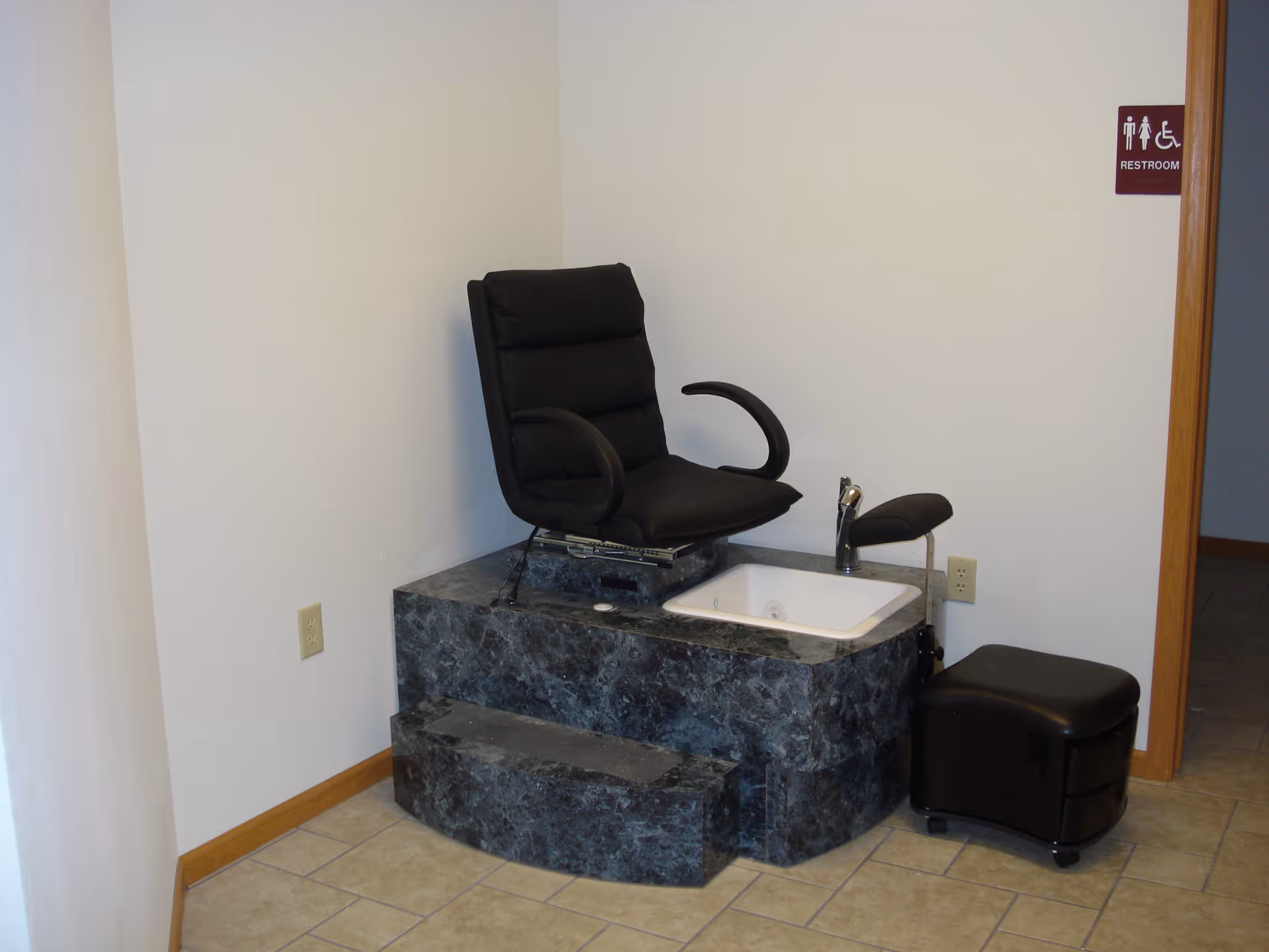 A pedicure station with a black cushioned chair mounted on a marble-like platform with a built-in foot bath. There is a small black stool on wheels next to the platform. The room has beige tiled flooring and white walls. A restroom sign is visible on the wall near a wooden door frame.