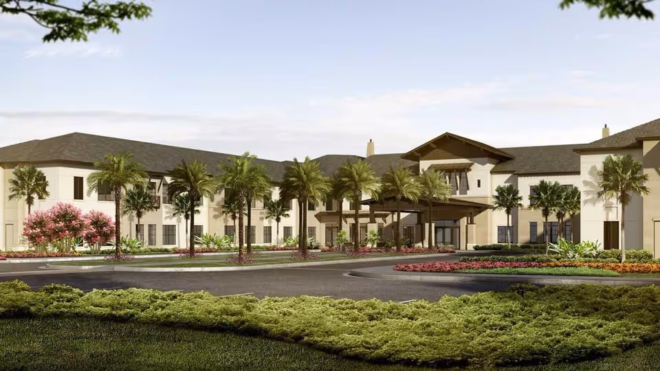 Front exterior of a two-story assisted living building with palm trees, landscaped gardens, and a circular driveway.