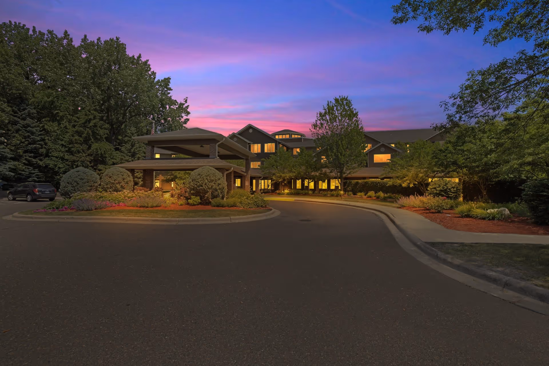 Exterior view of The Lodge at White Bear senior living facility at dusk, showing a large building with multiple windows lit from inside, surrounded by trees and landscaped bushes, under a purple and pink sky.