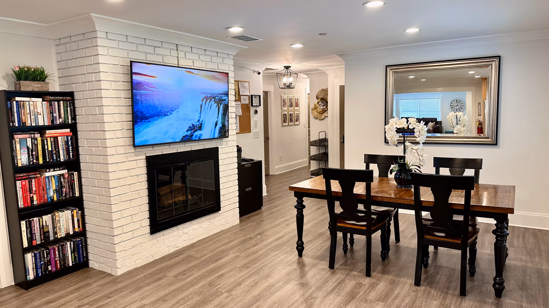 Dining area with a wooden table and chairs, a wall-mounted TV above a white brick fireplace, a bookshelf and a large mirror.