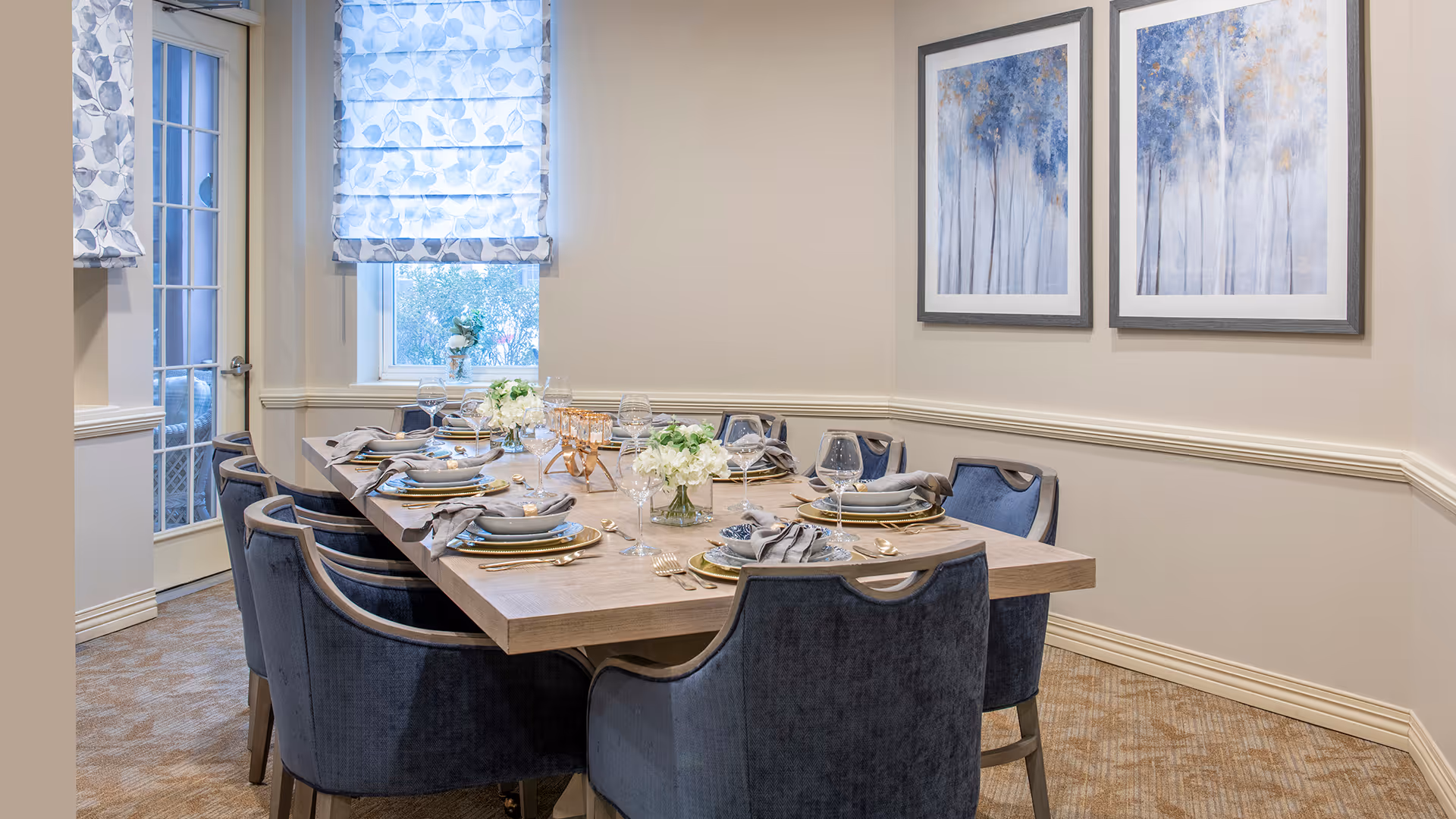 A well-lit dining room with a rectangular wooden table set for eight people. The table is arranged with plates, napkins, glasses, and floral centerpieces. There are eight upholstered chairs around the table. The room has beige walls with white trim, two framed artworks featuring blue and gold trees, a window with patterned blinds, and a glass door.