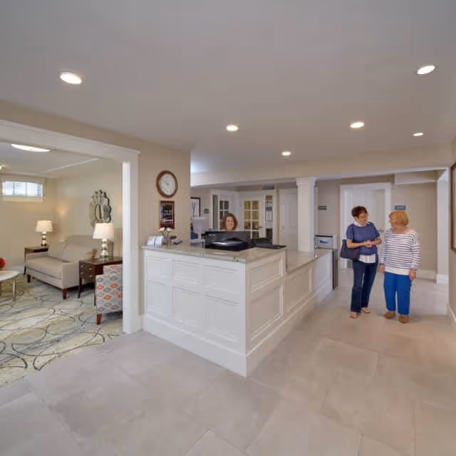 Interior view of a senior living facility reception area with a white reception desk, a staff member behind the desk, and two elderly women walking and talking nearby. To the left, there is a cozy sitting area with a sofa, armchair, side tables, lamps, and a decorative mirror on the wall.